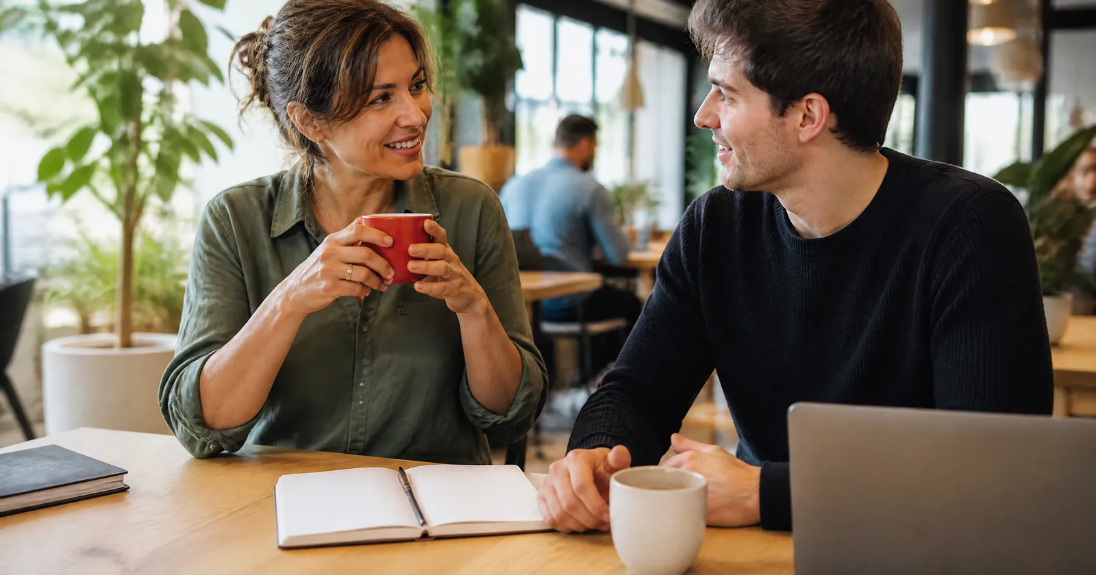 A business owner holding a red mug talks with a young web designer at a café table in bright daylight.