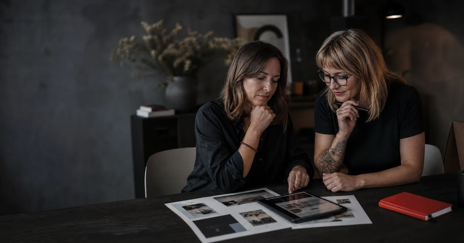 A business owner and a web designer review printed website layouts and a tablet at a dark table, a red notebook beside them.
