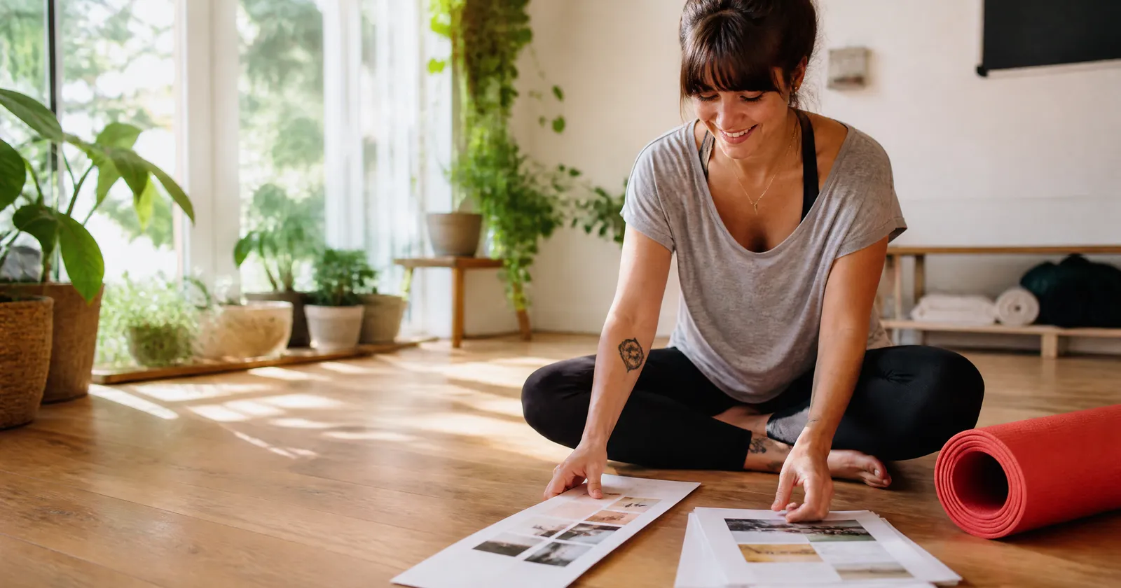 A smiling yoga studio owner lays out two printed page layouts on the floor of her sunny studio, a red mat beside her.