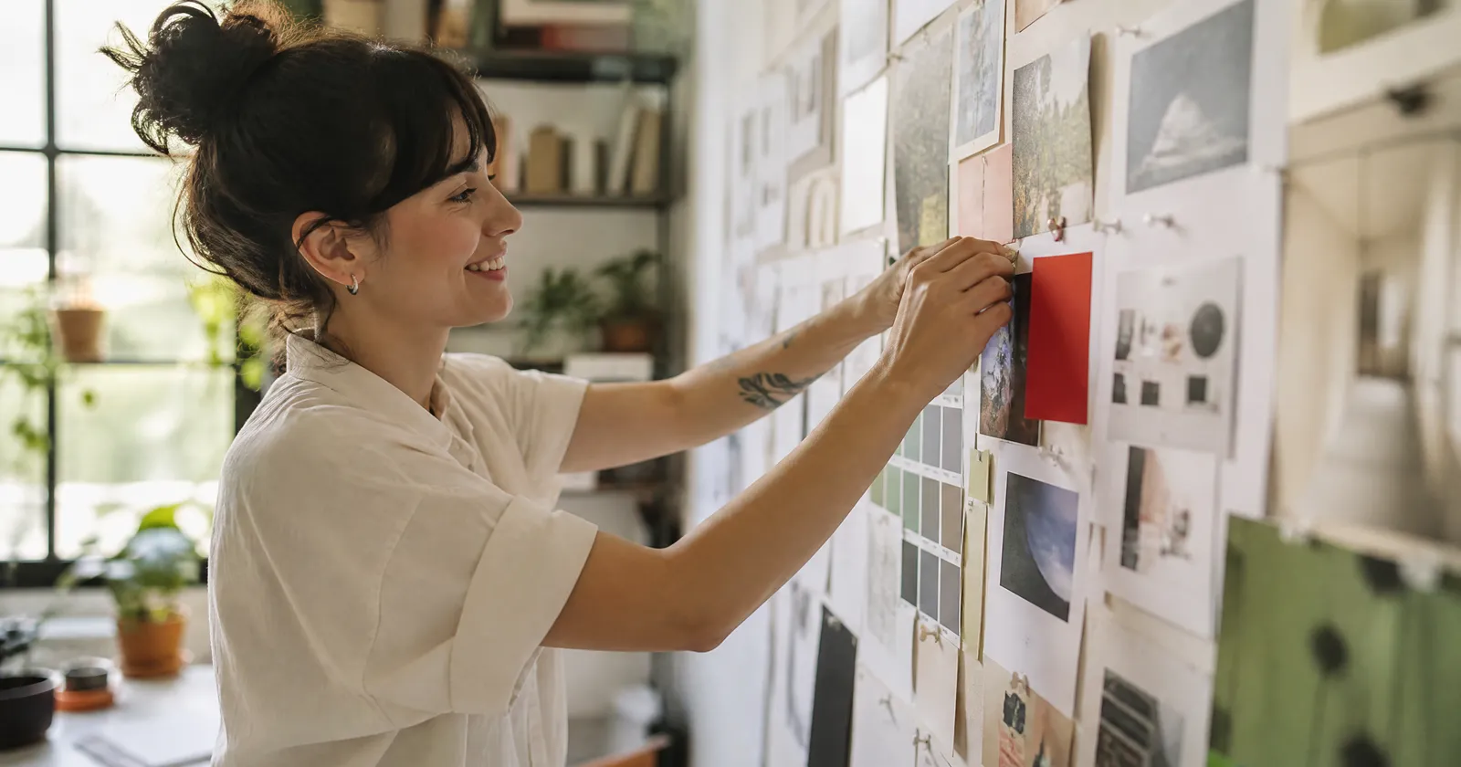 A smiling designer pins website prints, photos and a red colour swatch onto a mood board in a sunny studio.